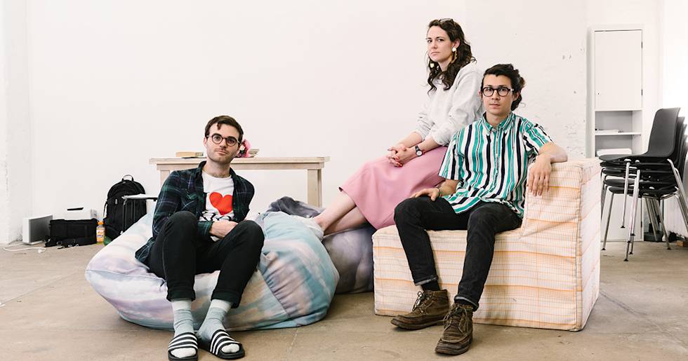 Three young professionals relaxing in casual startup office space with bean bag chair and minimalist furniture against white wall