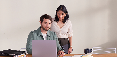Two colleagues working together at a desk with a laptop, papers, and office supplies against a neutral background