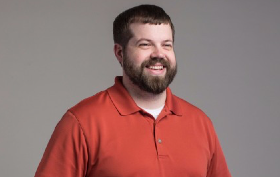 Professional headshot of a smiling person with a full beard wearing a red polo shirt against a gray background