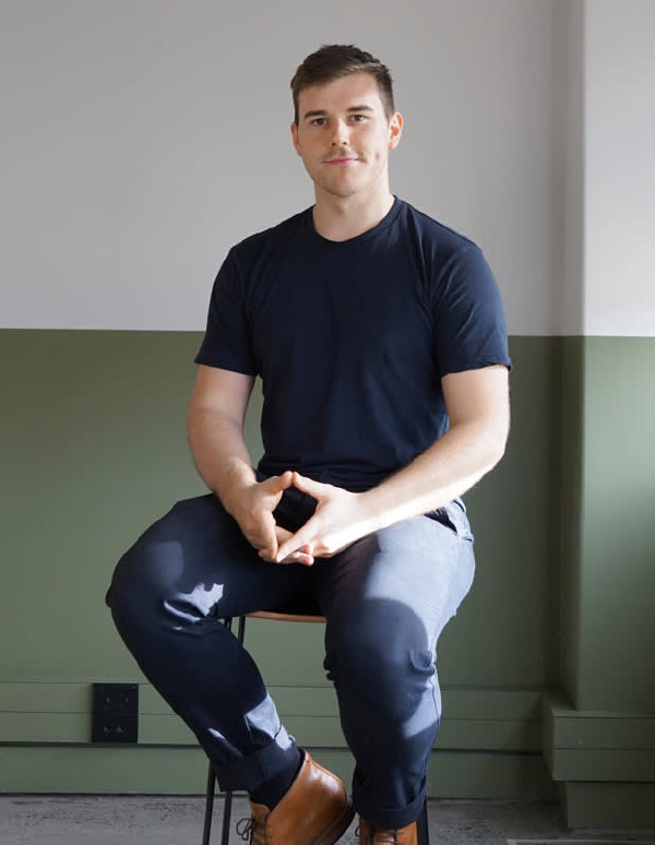 A person in a navy t-shirt and dark pants sits on a chair against a two-toned wall, wearing brown leather boots.