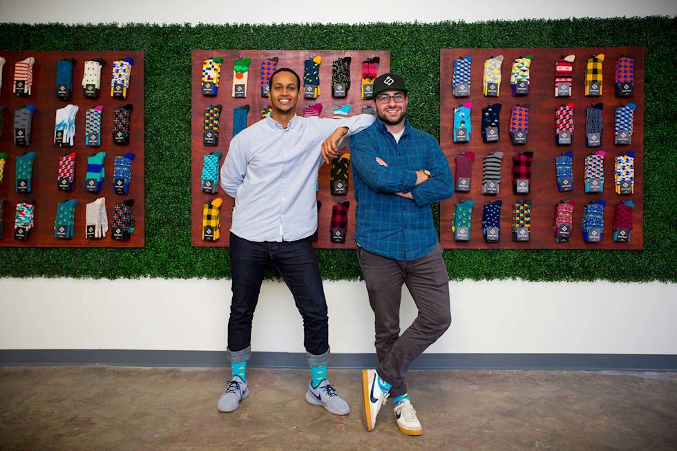 Stefan Lewinger and Futhum Tewolde of Sock Fancy smiling in front of a wall covered in socks. 