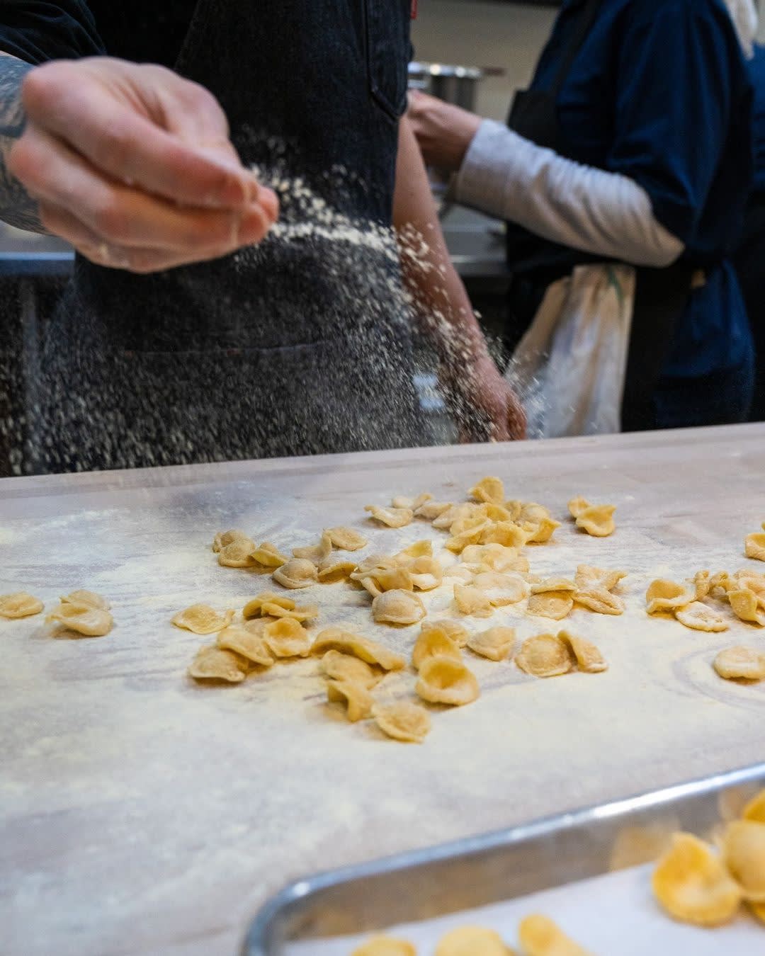 A chef's hand is shown sprinkling flour over a table with handmade, uncooked orecchiette pasta
