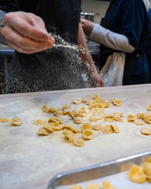 A chef's hand is shown sprinkling flour over a table with handmade, uncooked orecchiette pasta