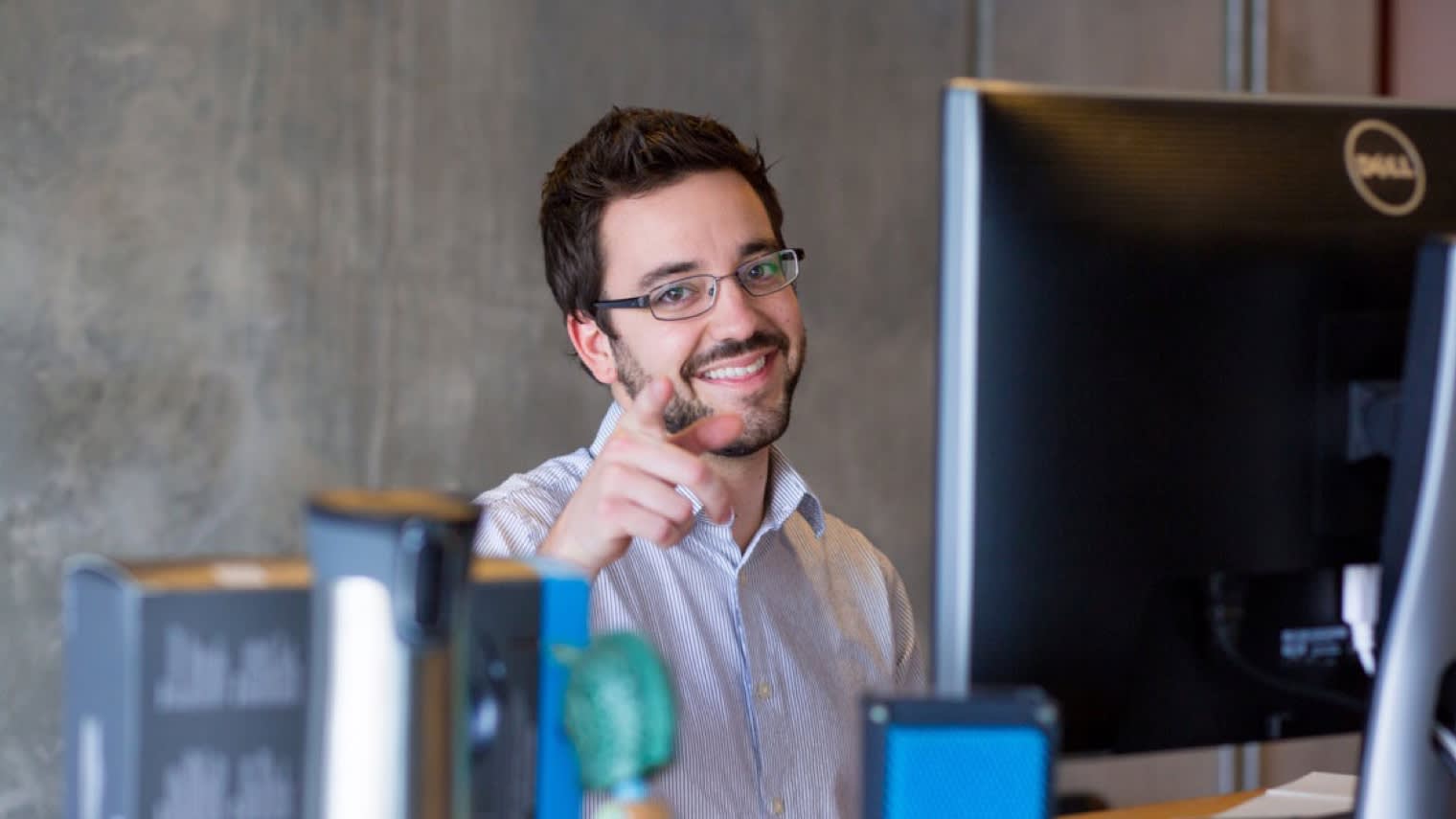 Professional in glasses working at desk with Dell monitor, smiling and pointing while seated in modern office setting