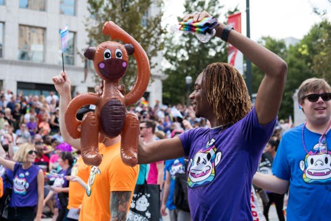 Festive parade crowd with person holding inflatable monkey balloon, wearing purple shirt with cartoon character design
