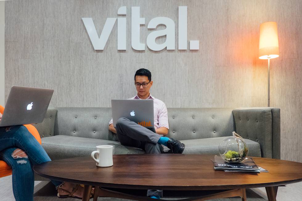 Modern office space with "vital" logo on wall, featuring grey couch, round wooden table, and MacBook users in casual work setting