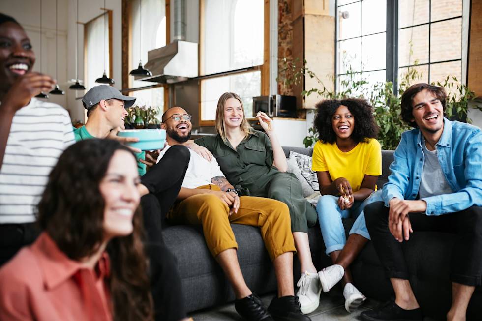 Diverse group of friends laughing together on a gray couch in a bright, modern room with plants and large windows