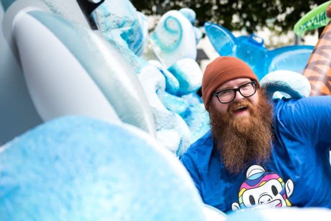 Bearded person in blue jacket and orange beanie smiles while lounging in a pile of large inflatable pool toys and floats