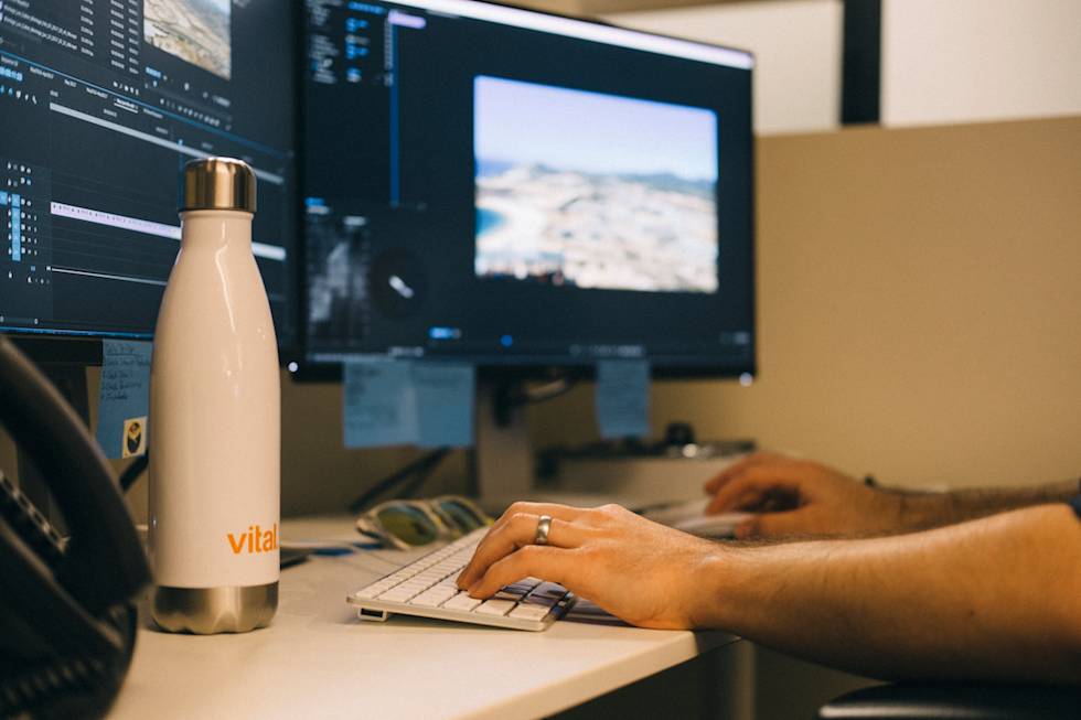 Video editor working at desk with beige Vital water bottle, keyboard, and dual monitors showing editing software interface