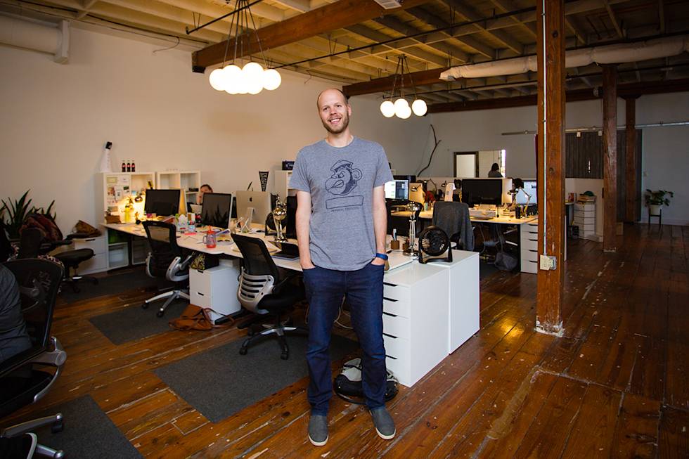 Modern open office space with wooden floors, exposed beams, globe lights, and white desks arranged in a collaborative workspace layout