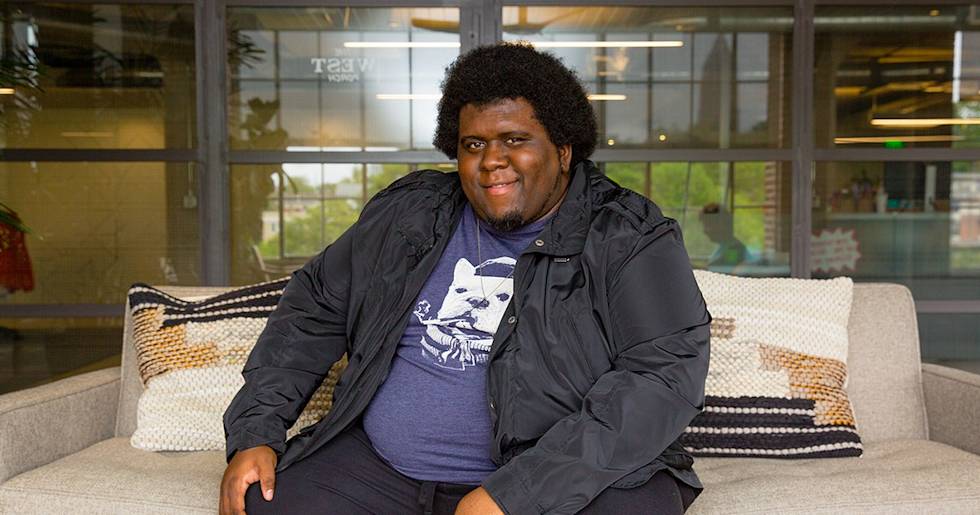 A person with an afro hairstyle sits smiling on a gray couch, wearing a navy graphic t-shirt and black jacket in a modern office setting