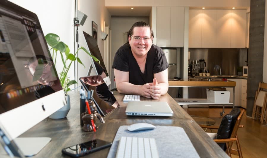 Person in black shirt smiling at desk with Apple computer setup, houseplant, and modern kitchen visible in background
