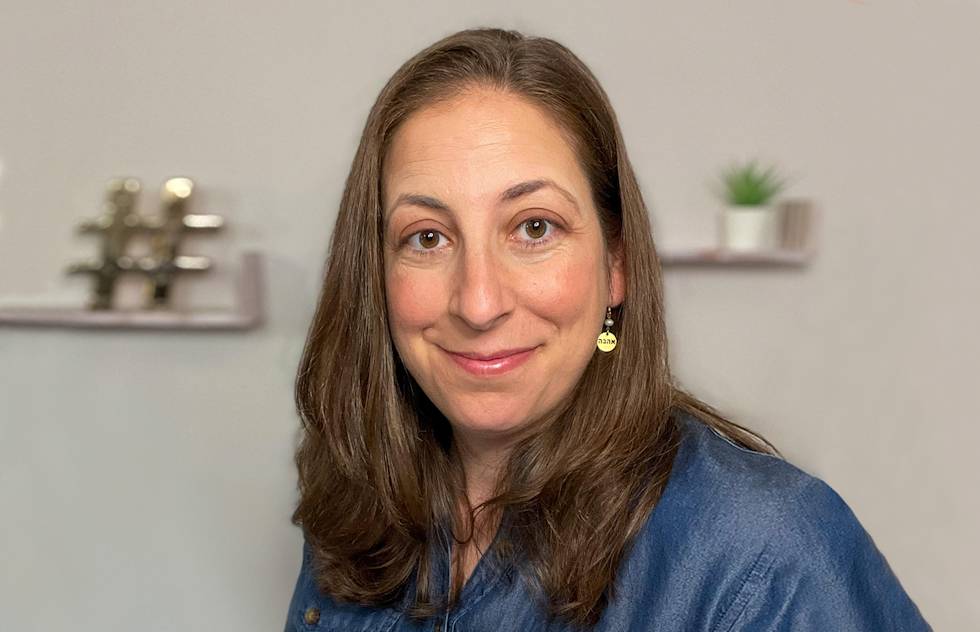 A warm smile and gentle expression on a person wearing a blue denim shirt, with decorative shelves and plants visible in the background.