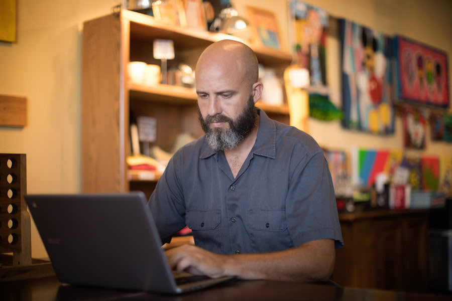 Professional with beard working on laptop in creative office space with colorful artwork and wooden shelving in background
