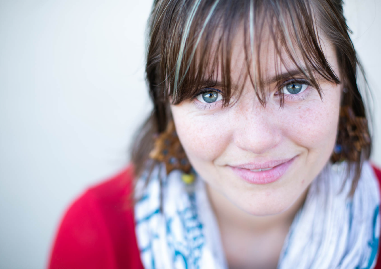 Close-up portrait of someone wearing a red top and patterned white scarf, with bangs and bright blue eyes, smiling warmly at camera