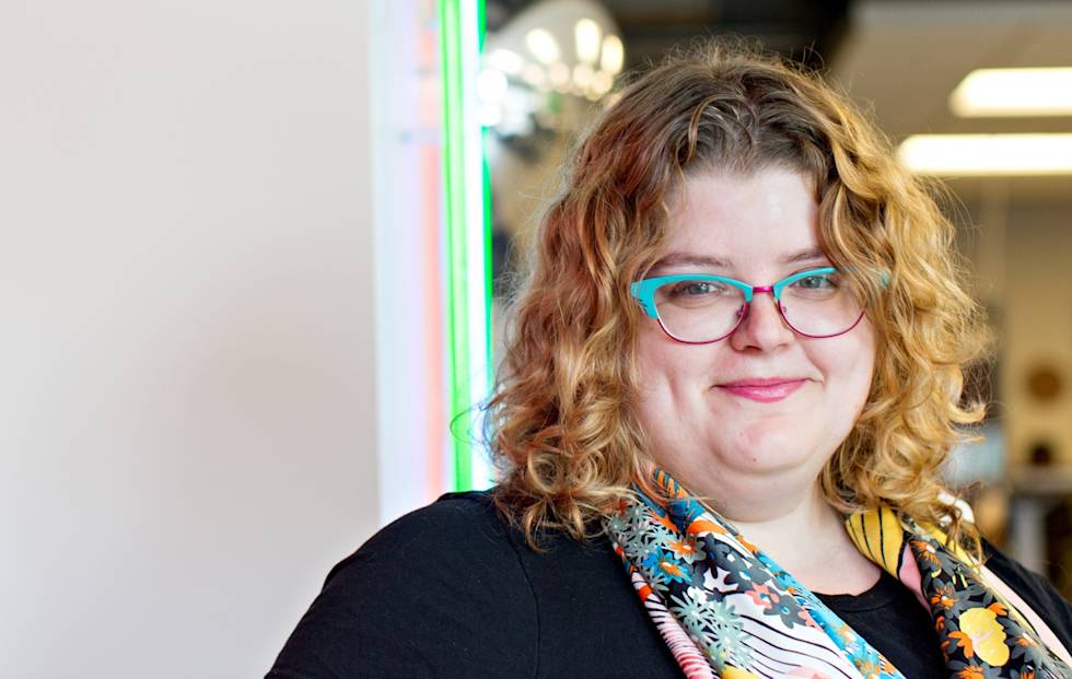 A smiling person with curly blonde hair wearing bright turquoise and red glasses and a black top with colorful floral pattern