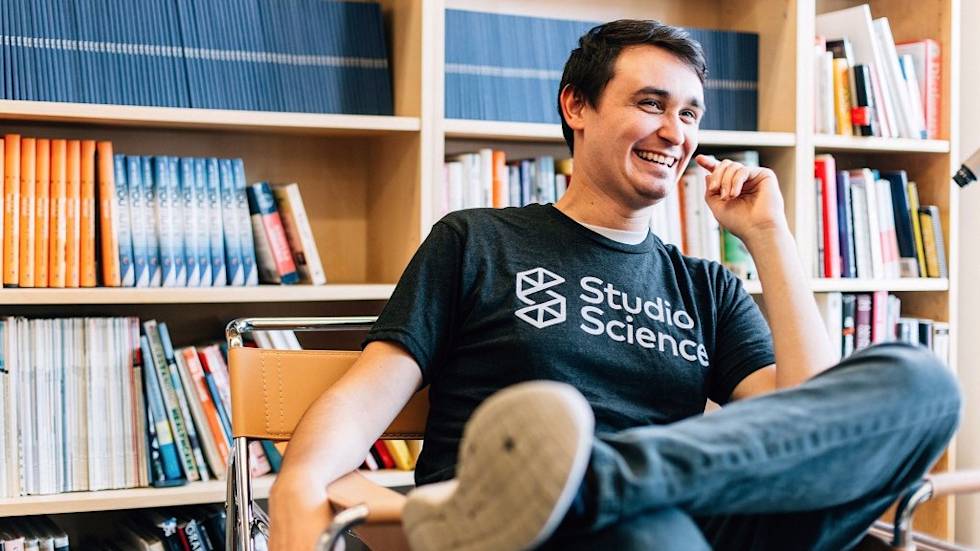 A person in a Studio Science t-shirt sits casually in a chair, smiling in front of bookshelves filled with colorful books