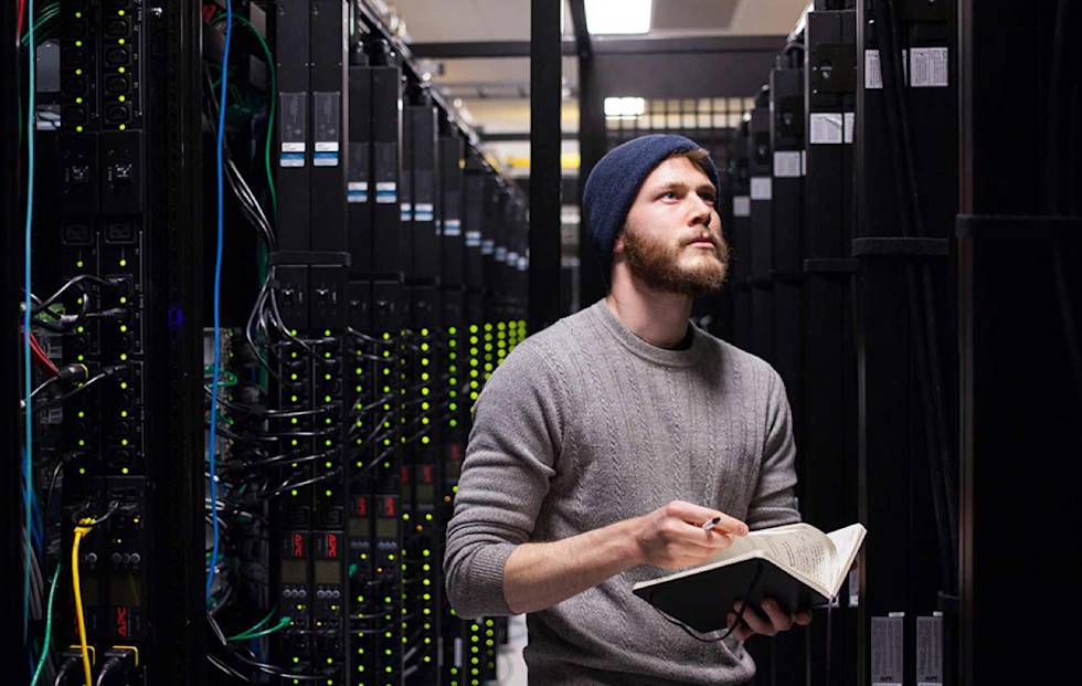 IT technician in beanie and sweater examining notes while standing in server room with glowing equipment racks