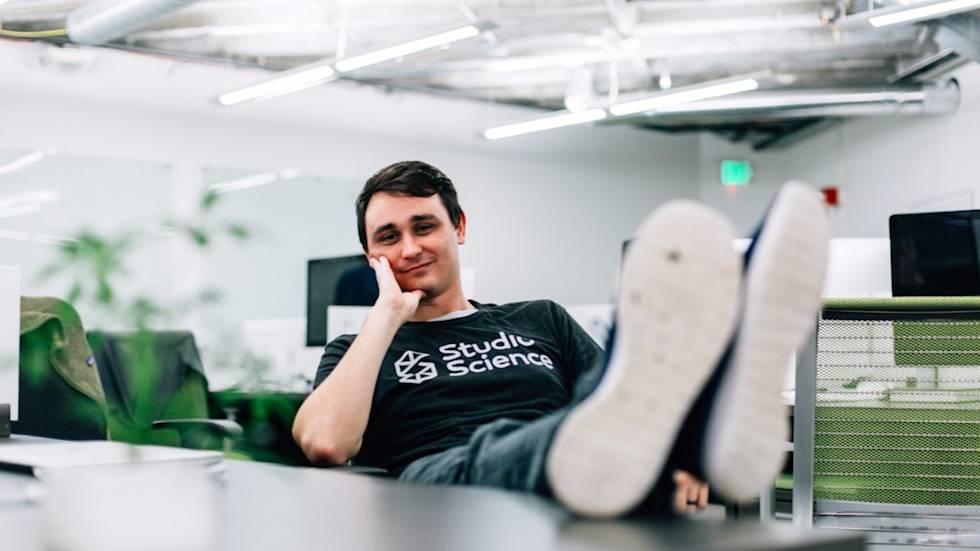 Relaxed employee in Studio Science t-shirt reclines at desk with feet up in modern office space with plants and fluorescent lighting