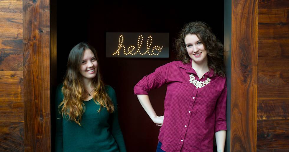Two smiling women stand in a doorway beneath an illuminated "hello" sign, one wearing green and one in burgundy with a statement necklace