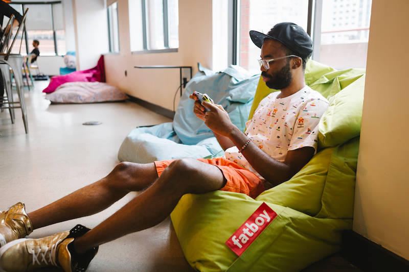 Person relaxing on green Fatboy beanbag chair in casual office space, using smartphone while wearing baseball cap and patterned shirt