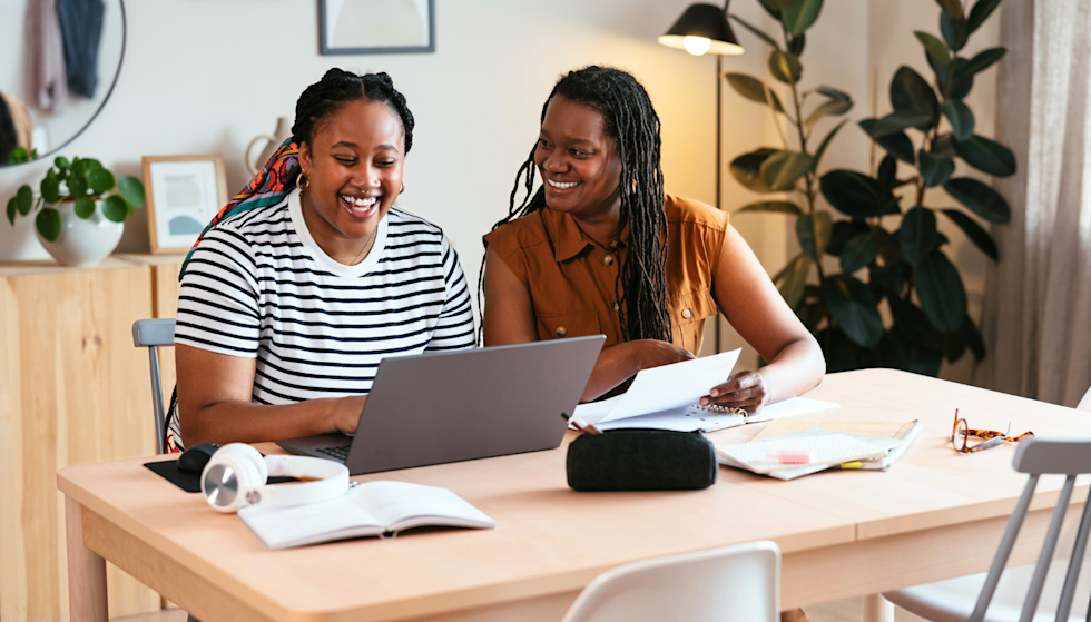 A cheerful teenager and her mother sitting together at a desk, looking at a laptop.