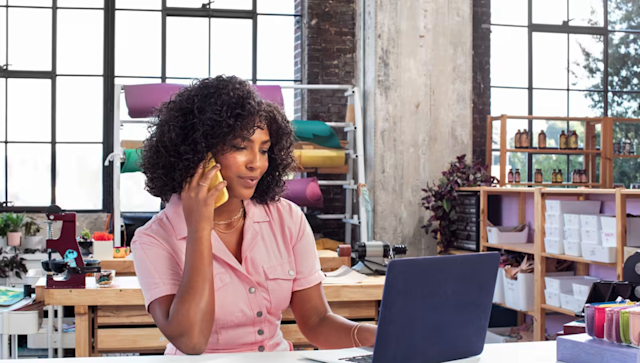 A woman with curly hair wearing a pink button-up shirt is sitting at a desk in a bright, modern studio. She is talking on a yellow phone while working on a laptop. Behind her are shelves with colorful fabric rolls, organized boxes, bottles, and plants, suggesting a creative or small business workspace.