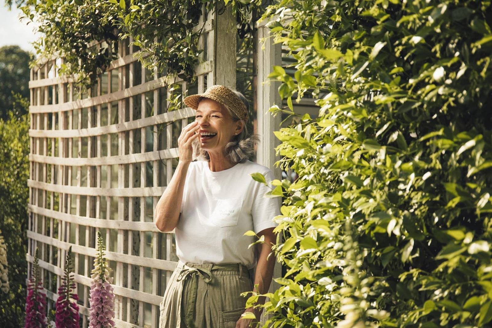 Woman in straw hat and white t-shirt laughing in sunny garden with white trellis and pink foxgloves