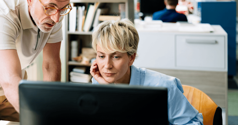 A woman with blond hair sits at a desk, her hand under her chin, as she and a man to her left, who is wearing glasses, both look at a computer screen.