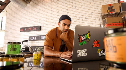 Person working on laptop at coffee shop counter with Oat Lord branded signage and merchandise visible in background