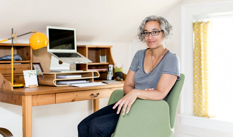 Professional in striped shirt sits at wooden desk with laptop, yellow lamp, and green chair in bright home office with yellow curtains