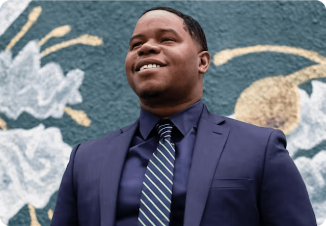 Image of Dundalan Sain, Director of Franchise Support, dressed in a suit and standing in front of a colorful flower mural painted on a wall