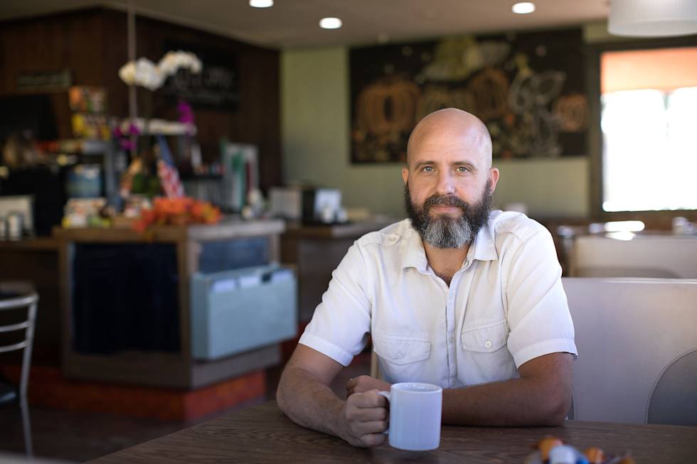 Professional in white shirt with full beard sits at wooden table in casual restaurant setting, holding a clear glass