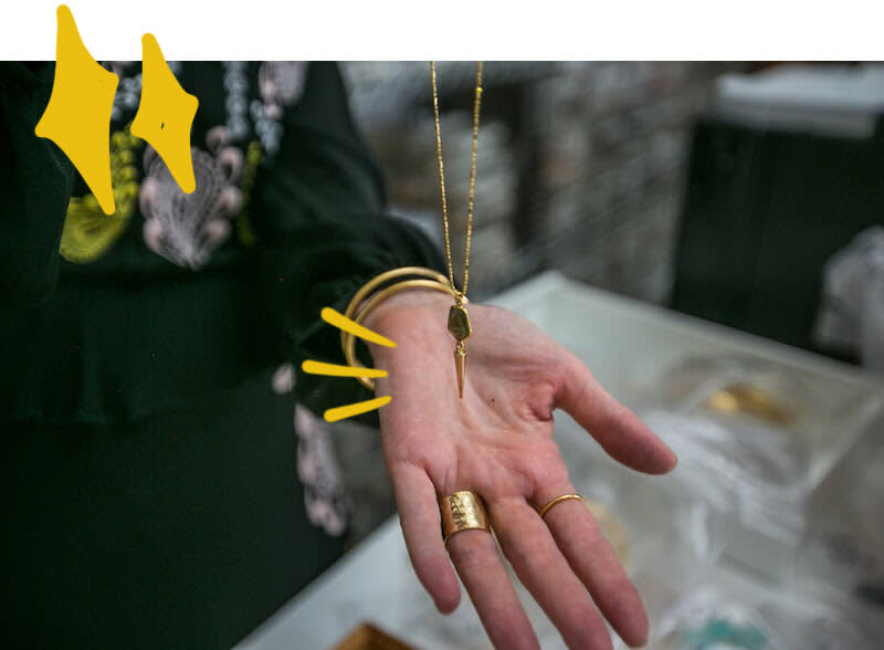 Hand wearing gold jewelry displays a delicate key necklace and textured ring against dark green background