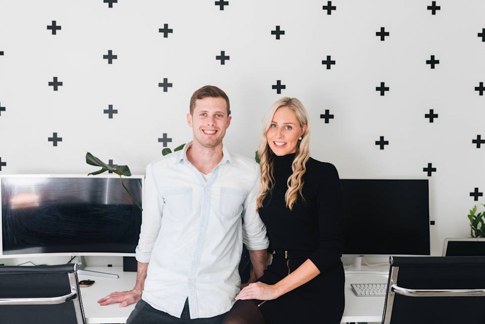 Two professionals in modern office with black plus sign pattern wall, white and black attire, standing between computer monitors