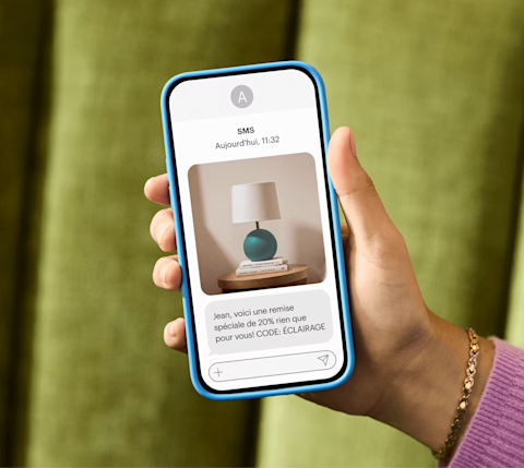 Une main tient un smartphone qui vient de recevoir un SMS marketing : « Alex, voici une réduction spéciale de 20 % rien que pour vous ! CODE : LUMINAIRE »
