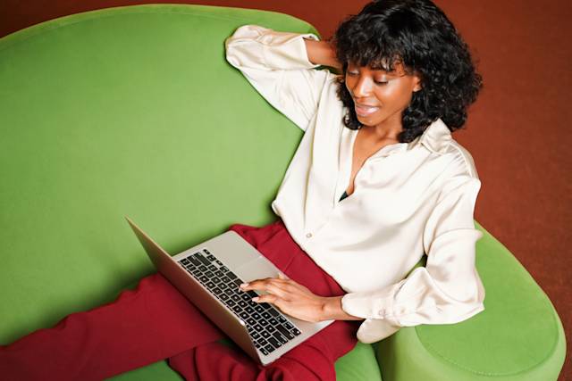 Femme en chemisier blanc travaillant sur un ordinateur portable tout en se relaxant sur une chaise vert vif, vêtue d'un pantalon bordeaux