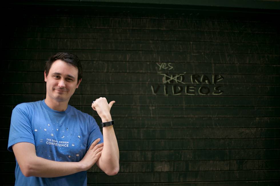 Person in blue conference t-shirt gestures toward graffiti reading "videos" on dark green wall