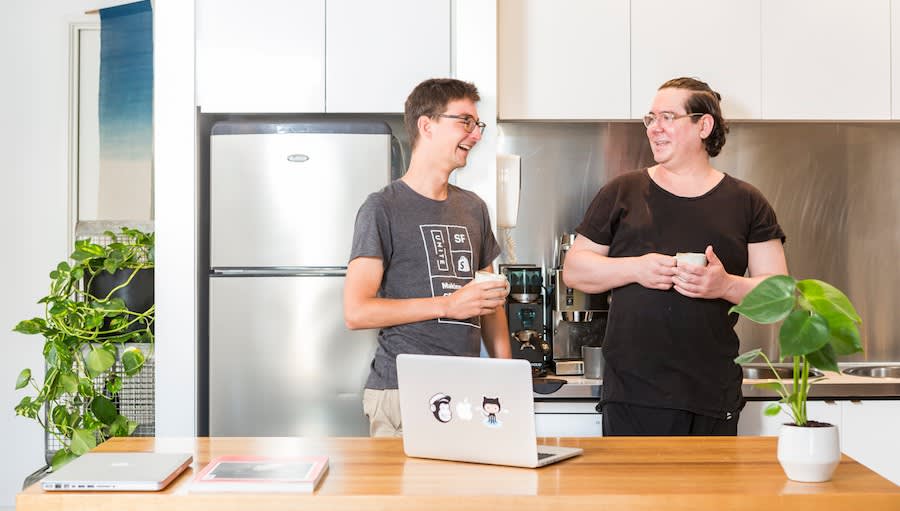 Two colleagues sharing coffee and conversation in a modern kitchen office space with laptops and plants on wooden desk