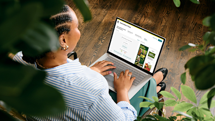 Person in striped shirt working on laptop surrounded by houseplants, viewed from above on wooden floor