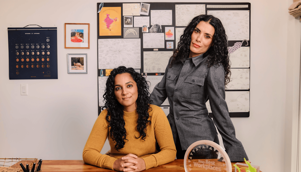 The CHANI co-founders pose in their office. Sonya Passi sits at the desk, and Chani Nicholas stands behind her with her hand on the back of the chair. In the center of the desk is a "2024 Inc. Best Workplaces" award.