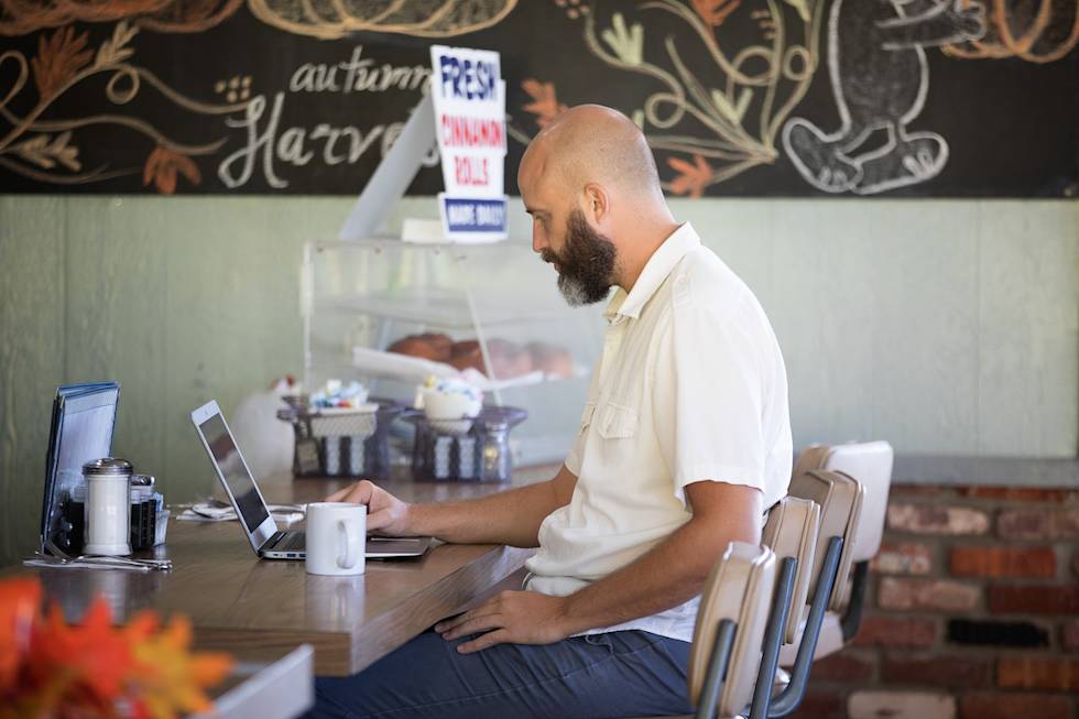 Bearded man in white shirt works on laptop at cafe counter with autumn-themed chalkboard art visible in background