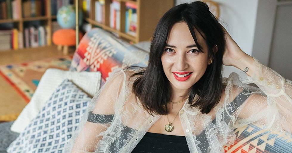A person in a sheer sparkly jacket smiles warmly while relaxing on patterned cushions, with bookshelves visible in the background