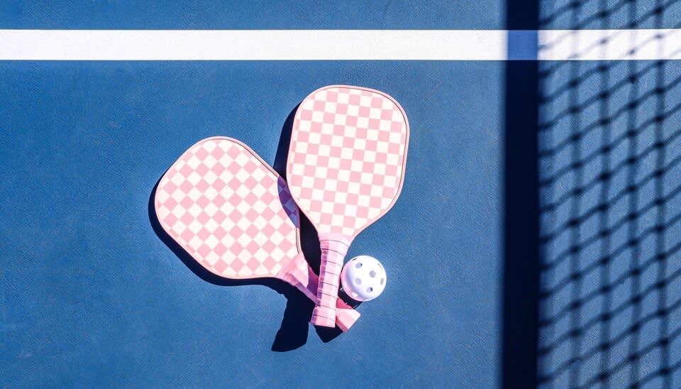 ​​Two pickleball paddles with a pink and white checkerboard pattern lay crossed on a blue pickleball court. A white pickleball sits next to them, and the shadow of a net is cast across the court to the right.]