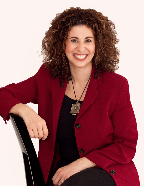 Professional headshot of a person with curly brown hair wearing a burgundy blazer and black top, smiling warmly against a light background