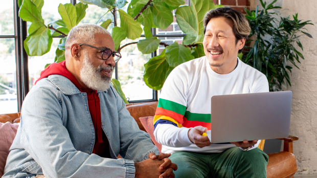 2 men sit on a couch in an office, while one is holding a laptop happily discussing their marketing strategy.