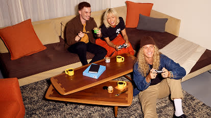 Three friends relaxing on a beige sectional sofa with orange pillows, drinking from yellow mugs around a triangular coffee table