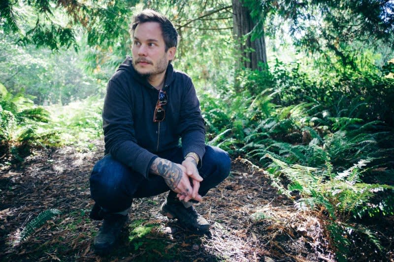 Person in navy hoodie crouching in lush forest setting surrounded by ferns and evergreen trees in dappled sunlight