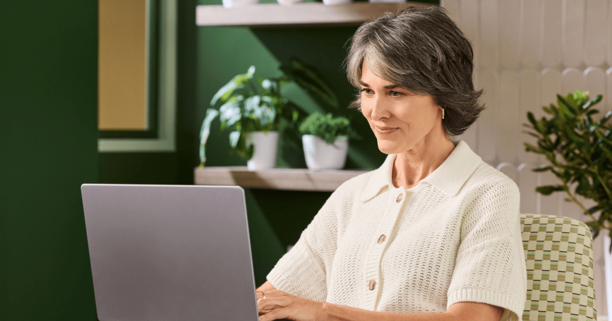 A woman, wearing a white sweater, ​​sits in a well-lit room with a calming green color scheme, using a laptop.