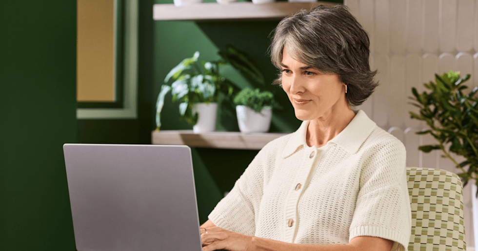 A woman, wearing a white sweater, ​​sits in a well-lit room with a calming green color scheme, using a laptop.